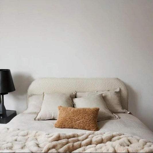 A minimalist bedroom with a Teddy Boucle Bed featuring a curved, beige upholstered headboard. The bed is dressed in light beige bedding, with three matching square pillows and a contrasting tan rectangular pillow. A black matte table lamp sits on a black bedside table, adding contrast to the calm, neutral-toned space. The plain white wall enhances the understated, soft-textured aesthetic.
