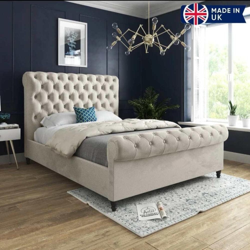 A stylish bedroom featuring the luxurious Toronto Scroll Bed with a high tufted headboard and footboard in light beige. Dressed in a cream duvet, gray blanket, and a blue and white geometric pillow, it rests on a patterned rug over warm wooden floors. A modern metallic chandelier hangs above dark blue walls, while potted plants add freshness. A "MADE IN UK" logo with the UK flag appears in the top right corner.