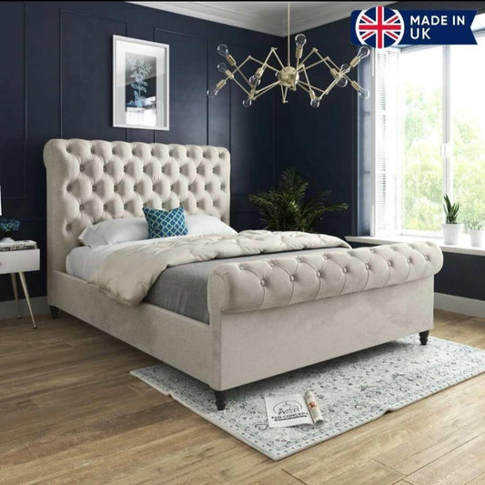A stylish bedroom featuring the luxurious Toronto Scroll Bed with a high tufted headboard and footboard in light beige. Dressed in a cream duvet, gray blanket, and a blue and white geometric pillow, it rests on a patterned rug over warm wooden floors. A modern metallic chandelier hangs above dark blue walls, while potted plants add freshness. A "MADE IN UK" logo with the UK flag appears in the top right corner.