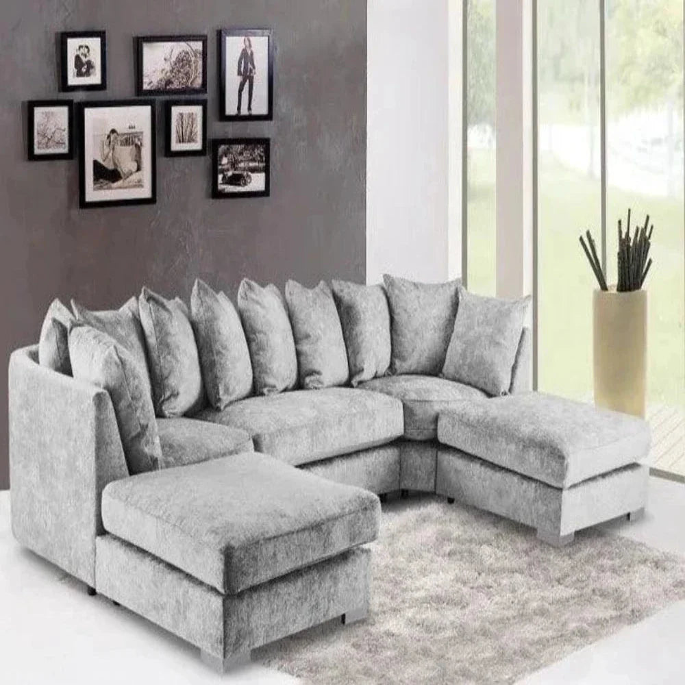 A modern living room features a U-shaped Elveden sofa in plush grey, adorned with matching cushions. It sits on a light grey textured rug. The dark grey wall showcases an asymmetrical collage of black-framed art, while large floor-to-ceiling windows offer views of greenery. A tall beige planter with decorative accents adds a natural touch to the airy space.