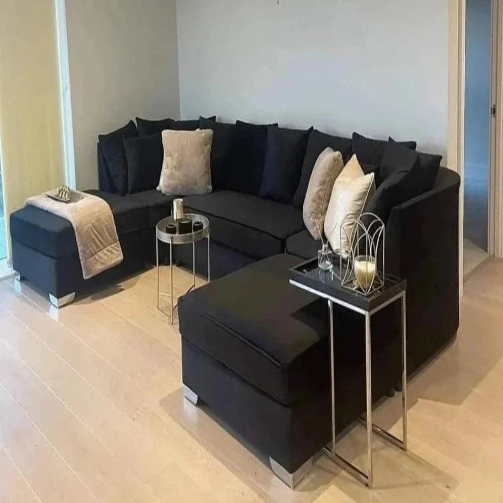 This modern living room showcases a U-shaped Elveden sofa in black, accented with large plush beige and cream cushions. A sleek round silver side table holds three candles, while a rectangular metallic side table next to the sofa features a decorative candle holder. A soft beige throw blanket drapes over the chaise section. Light hardwood flooring adds warmth to the space, which includes a doorway leading to another room.