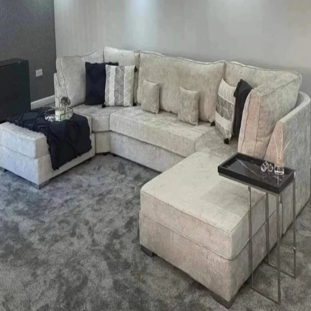 This modern living room features a U-shaped Elveden sofa upholstered in light beige fabric, accented with matching pillows in geometric patterns. A dark throw blanket is draped over one side. The gray carpet adds softness, while a small black side table with a metallic frame holds decorative items. Soft neutral gray walls enhance the room's contemporary feel, with a darker feature wall visible in the background.