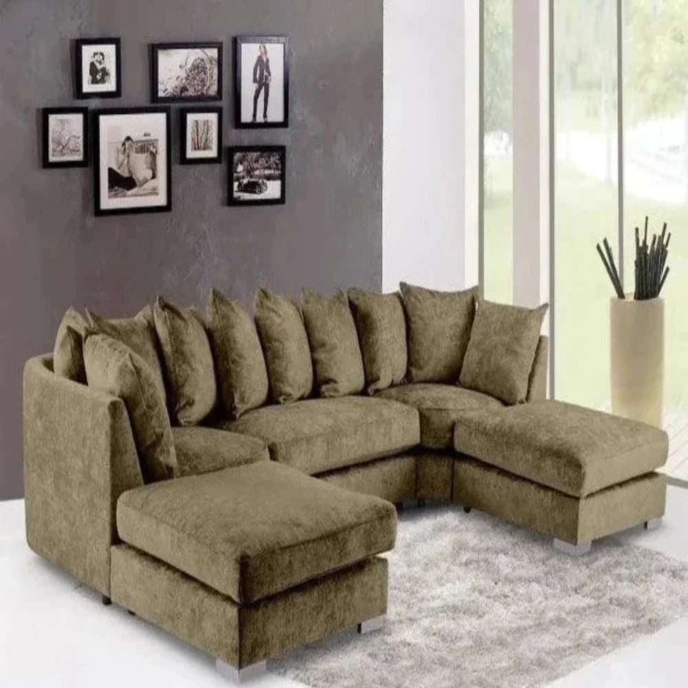 This contemporary living room features a U-shaped Elveden sofa upholstered in textured brown fabric, arranged for a cozy atmosphere with plush cushions. A soft light gray shag rug adds texture beneath the sofa. The solid gray back wall contrasts beautifully and is adorned with a cluster of framed black-and-white photographs. Floor-to-ceiling windows allow natural light to fill the room, revealing greenery outside, while a tall beige vase with decorative sticks enhances the decor.