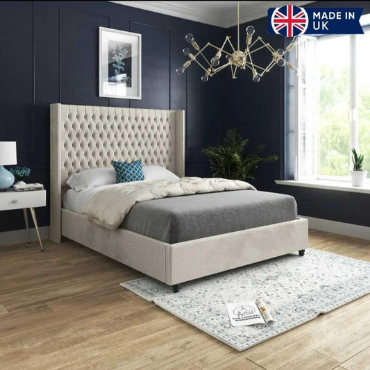 The image features a stylish bedroom with a Victoria Winged Imperial Bed, showcasing a tufted beige headboard. The bed is adorned with a light gray comforter, white sheets, and a decorative blue pillow. A white nightstand with a blue vase and lamp sits beside the bed. The deep navy walls are accented by a botanical print, while an exposed-bulb chandelier hangs above. A light-patterned rug covers the wood plank floor, and a green potted plant is positioned by the window, allowing filtered light in.