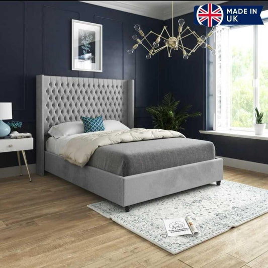 The image showcases a modern bedroom centered around a Victoria Winged Imperial Bed with a plush light gray tufted headboard. The bed features a gray comforter, white duvet, and a decorative blue pillow. A white bedside table with a blue vase and lamp sits beside it against a dark blue wall adorned with elegant molding and a plant print. A large window floods the room with natural light, highlighting a leafy green plant nearby.