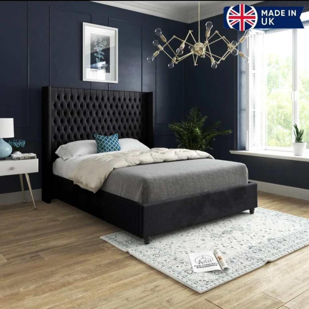 The image showcases a modern bedroom featuring a Victoria Winged Imperial Bed with a black tufted headboard, gray bedspread, white pillows, and a blue patterned cushion. The deep navy walls and gold geometric chandelier create a luxurious ambiance. A white side table with a blue lamp is positioned beside the bed. A tall indoor plant by a large window with white blinds allows natural light in. The light wooden floor is covered with a pale patterned rug.