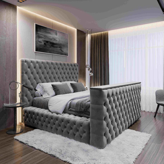 The image features a modern bedroom with a plush gray Walton TV Bed, showcasing a tufted headboard and footboard. The bed is layered with white and gray bedding, complemented by a monochromatic painting above it. A sleek round side table with a black lamp is positioned to the left. Sheer curtains over a large window allow soft light to fill the room, enhancing its muted tones, while a quilted gray chair adds elegance.