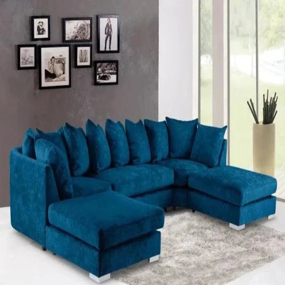 This modern living room showcases a U-shaped Elveden sofa upholstered in rich teal blue fabric, complemented by numerous matching pillows. It rests on a light gray fluffy area rug at the room's center. A dark gray textured wall displays a stylish arrangement of black-framed photographs. Floor-to-ceiling windows allow natural light to flood the space and reveal greenery outside, while a tall vase with long-stemmed plants adds a touch of nature.