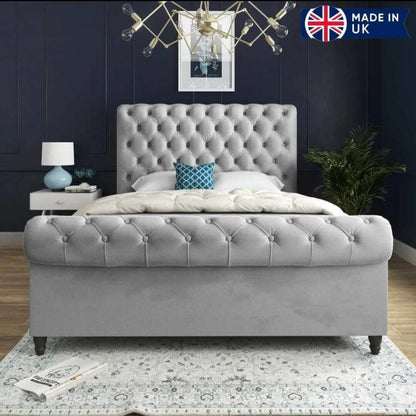 A modern bedroom showcases a plush Toronto Scroll Bed in gray, featuring a tufted headboard and footboard against a dark blue paneled wall. It's dressed with a white comforter and a blue geometric pillow for contrast. A contemporary light fixture with exposed bulbs hangs above. To the left, a white nightstand holds a blue lamp and books, while a leafy plant sits to the right. A light-colored patterned rug covers the wooden floor. In the top right corner, a banner with the Union Jack reads "MADE IN UK."