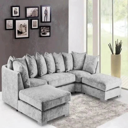 A modern living room features a U-shaped Elveden sofa in plush grey, adorned with matching cushions. It sits on a light grey textured rug. The dark grey wall showcases an asymmetrical collage of black-framed art, while large floor-to-ceiling windows offer views of greenery. A tall beige planter with decorative accents adds a natural touch to the airy space.
