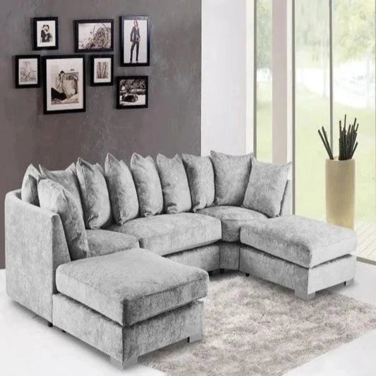 A modern living room features a U-shaped Elveden sofa in plush grey, adorned with matching cushions. It sits on a light grey textured rug. The dark grey wall showcases an asymmetrical collage of black-framed art, while large floor-to-ceiling windows offer views of greenery. A tall beige planter with decorative accents adds a natural touch to the airy space.