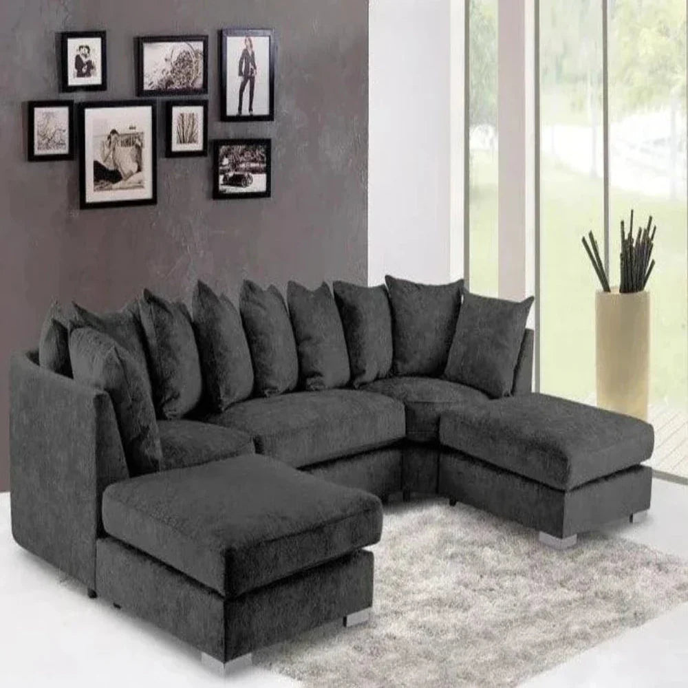 This modern living room features a spacious U-shaped Elveden sofa in dark gray with plush cushions, resting on a light gray textured rug. A dark wall behind the sofa displays a collage of seven black-framed pictures. Floor-to-ceiling windows provide natural light and views of greenery, while a tall beige planter with slender plants adds a touch of nature.