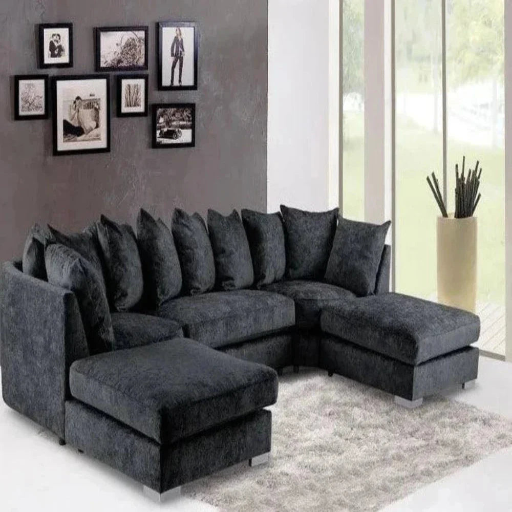 This modern living room features a U-shaped Elveden sofa in dark gray with a plush velvet texture and a chaise lounge. It sits on a light fluffy rug, complemented by a gray wall displaying a gallery of black-framed monochrome art. Floor-to-ceiling windows let in natural light and offer views of green grass, while a tall beige vase with decorative sticks adds a natural touch.